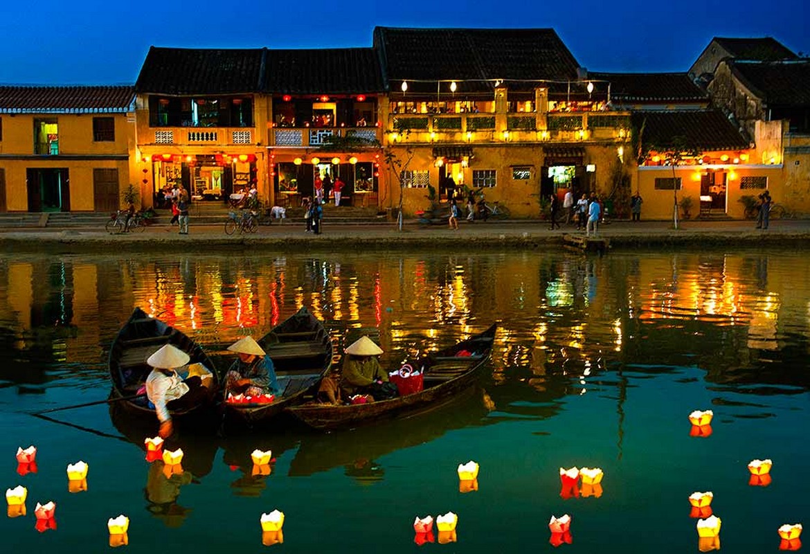 A lively riverside at night features traditional Vietnamese boats with travelers wearing conical hats, illuminated floating lanterns, and a yellow-colored building with balconies and red lanterns reflecting bright lights on the water.